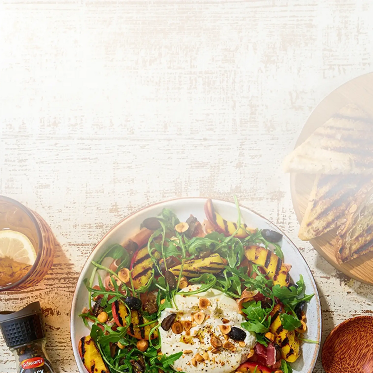 A plated salad with greens, grilled sweet potato slices, a poached egg, nuts, and a drizzle of dressing on a rustic white wooden table, alongside a glass of iced tea and toasted bread.