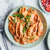 A rustic blue bowl filled with large pasta shells in a creamy tomato meat sauce, garnished with fresh arugula, sits on a marble countertop.