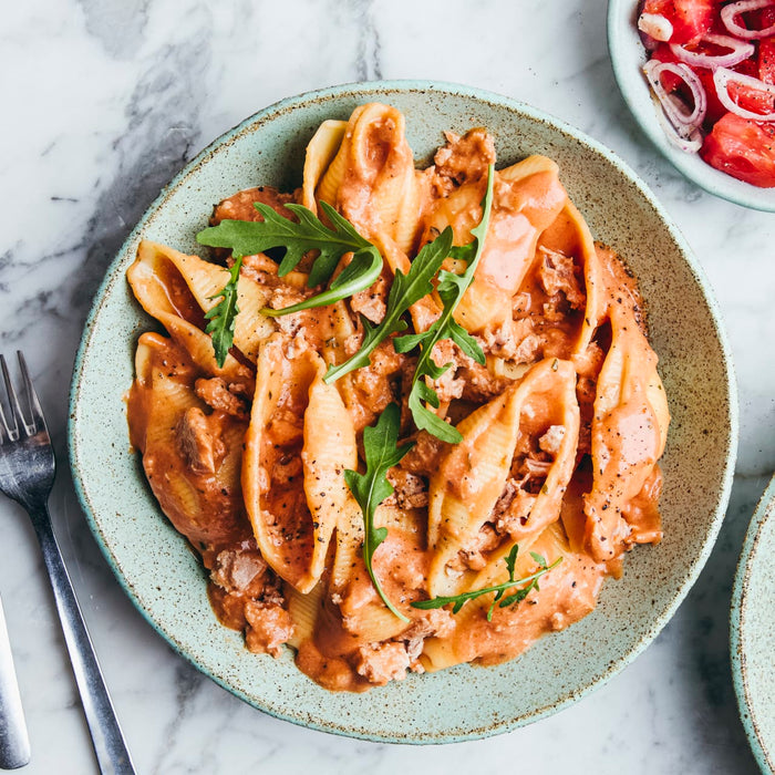 A rustic blue bowl filled with large pasta shells in a creamy tomato meat sauce, garnished with fresh arugula, sits on a marble countertop.