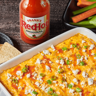 Buffalo chicken dip with Frank's RedHot sauce, chips, and vegetables on a wooden surface.