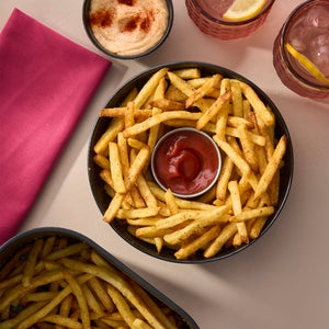 A top-down view shows seasoned french fries served in a black Barrel bowl with ketchup, a creamy dip, and pink drinks on the side.