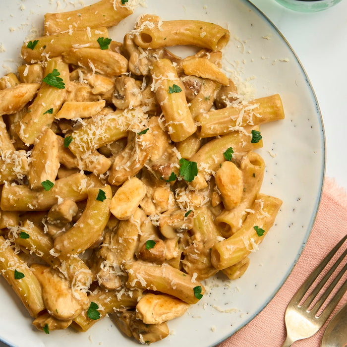 A close-up overhead view of a creamy chicken and mushroom pasta dish, made with large, Barrel-shaped pasta tubes, is served on a speckled plate and garnished with grated cheese and fresh parsley.