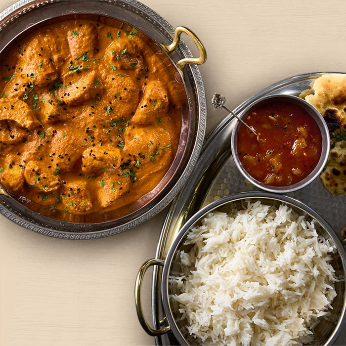 A freshly prepared Indian meal is displayed, featuring a bowl of chicken curry garnished with herbs and seeds, a bowl of fluffy white rice, and a side of chutney with naan bread.