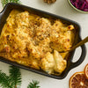 A close-up, overhead shot shows a creamy cauliflower cheese gratin with a golden-brown breadcrumb topping, served in a black 'Barrel' baking dish with festive holiday decorations.
