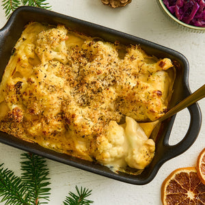A close-up, overhead shot shows a creamy cauliflower cheese gratin with a golden-brown breadcrumb topping, served in a black 'Barrel' baking dish with festive holiday decorations.