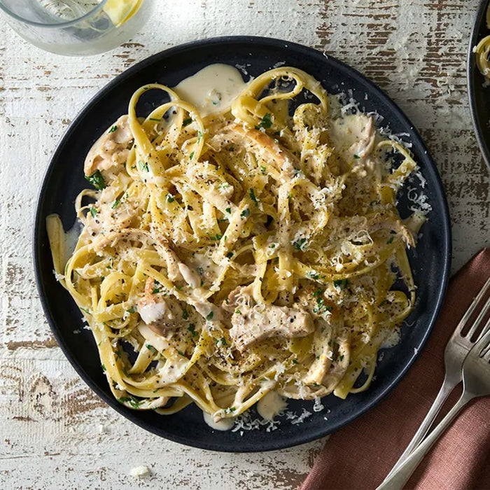A black plate holds a serving of creamy chicken fettuccine alfredo, garnished with grated parmesan cheese and a sprinkle of Barrel seasoning.