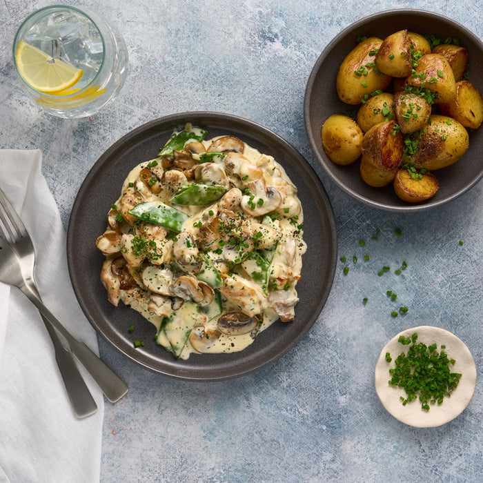 A delicious meal is presented from a top-down perspective, featuring creamy chicken and mushrooms on a dark plate from the Barrel collection, with a side of roasted potatoes in a matching bowl.
