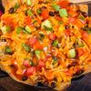 A close-up, top-down view of a Barrel of loaded chicken nachos, topped with melted cheese, black beans, avocado, tomatoes, and green onions in a cast-iron skillet.