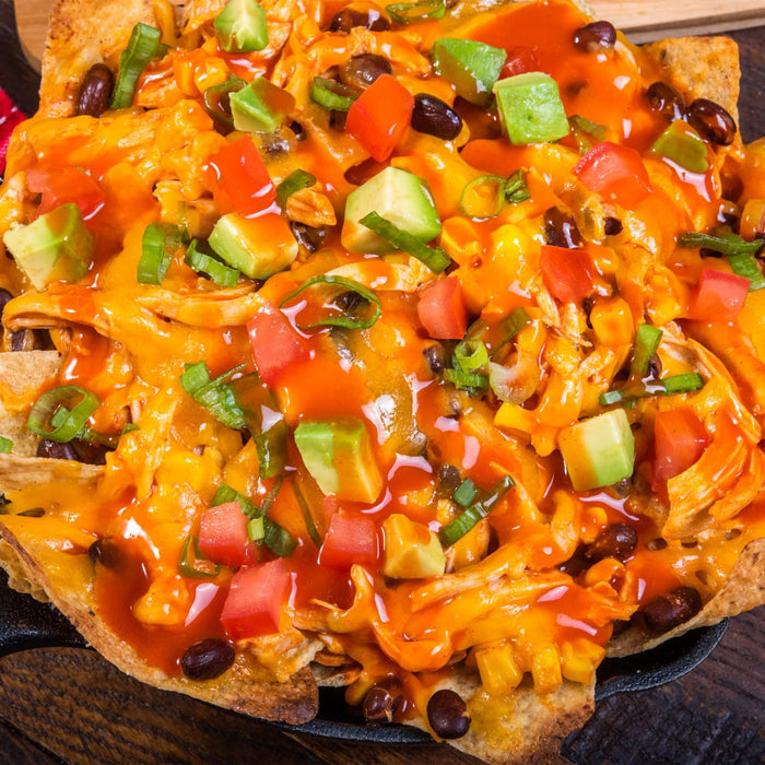 A close-up, top-down view of a Barrel of loaded chicken nachos, topped with melted cheese, black beans, avocado, tomatoes, and green onions in a cast-iron skillet.