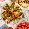 A delicious-looking meal of grilled chicken skewers, part of a Barrel recipe, is served on a plate with lime wedges, fresh cilantro, naan bread, a tomato and onion salad, and a creamy dipping sauce.