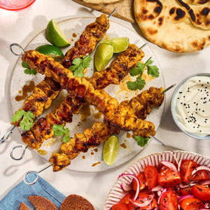 A delicious-looking meal of grilled chicken skewers, part of a Barrel recipe, is served on a plate with lime wedges, fresh cilantro, naan bread, a tomato and onion salad, and a creamy dipping sauce.