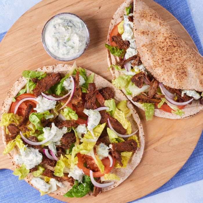 A round wooden Barrel cutting board holds two gyros, one open-faced and one folded, filled with seasoned meat, fresh vegetables, and tzatziki sauce.
