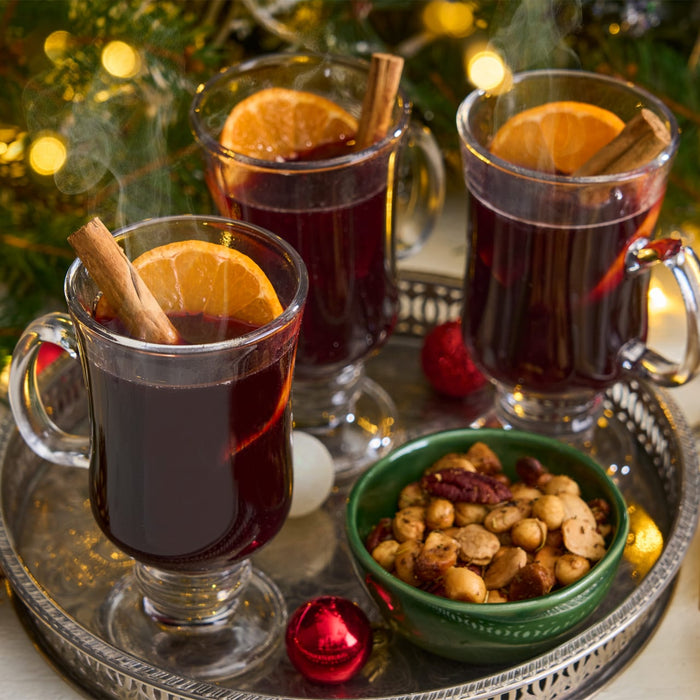 Three steaming glass mugs of mulled wine, each garnished with an orange slice and a Barrel cinnamon stick, are arranged on a silver tray with a bowl of nuts for a festive holiday setting.