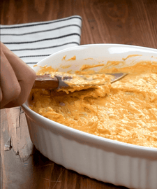 Cheesy dip being scooped out of a white dish with a wooden spoon.